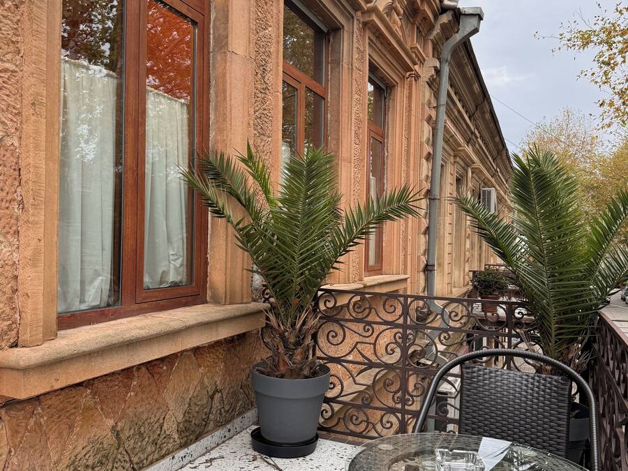 Un área de estar al aire libre con una mesa de vidrio y sillas de mimbre, una planta en maceta y un edificio con ventanas amplias y muros de piedra.