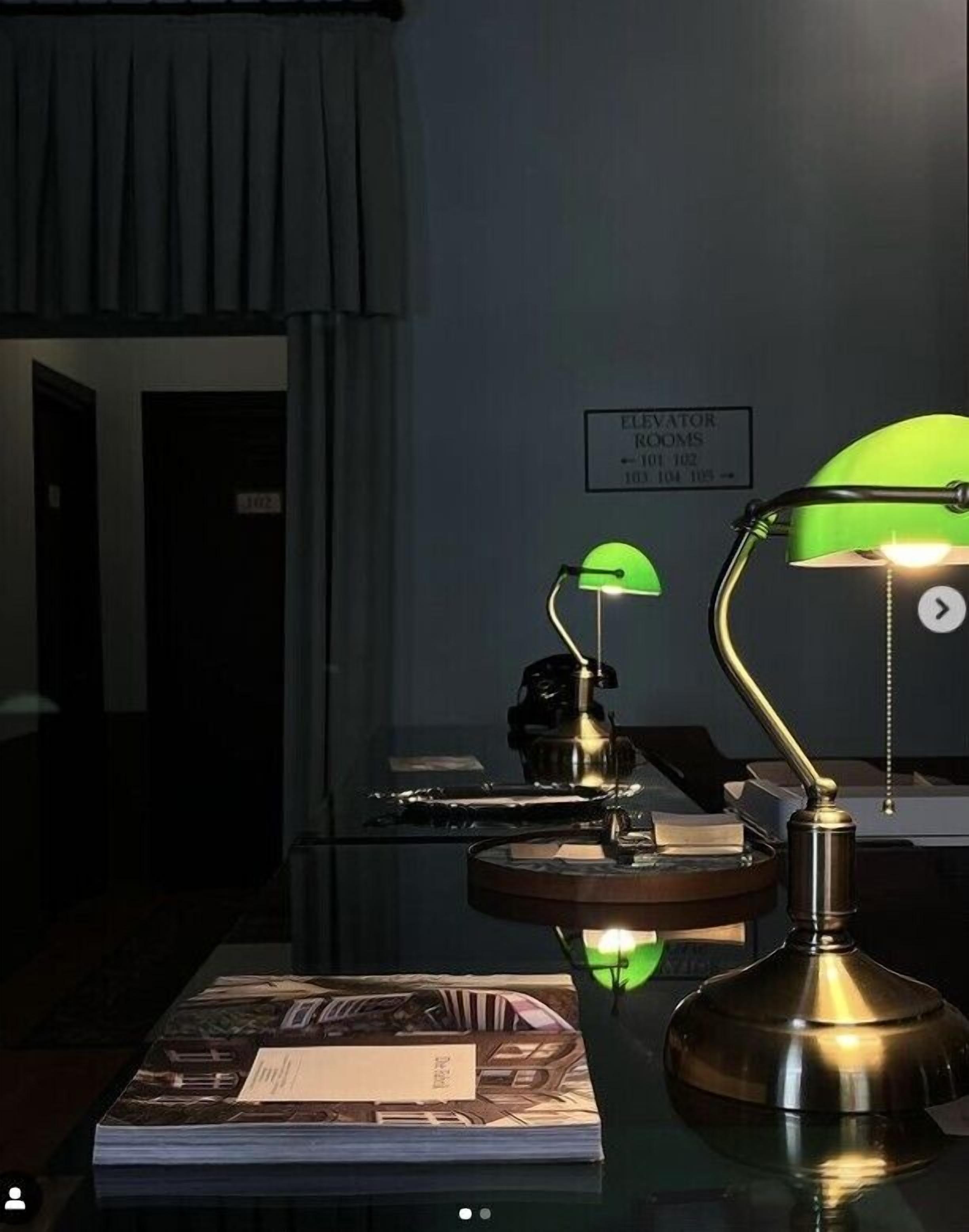 A desk with two green desk lamps, a book, and a sign indicating elevator rooms on the wall.