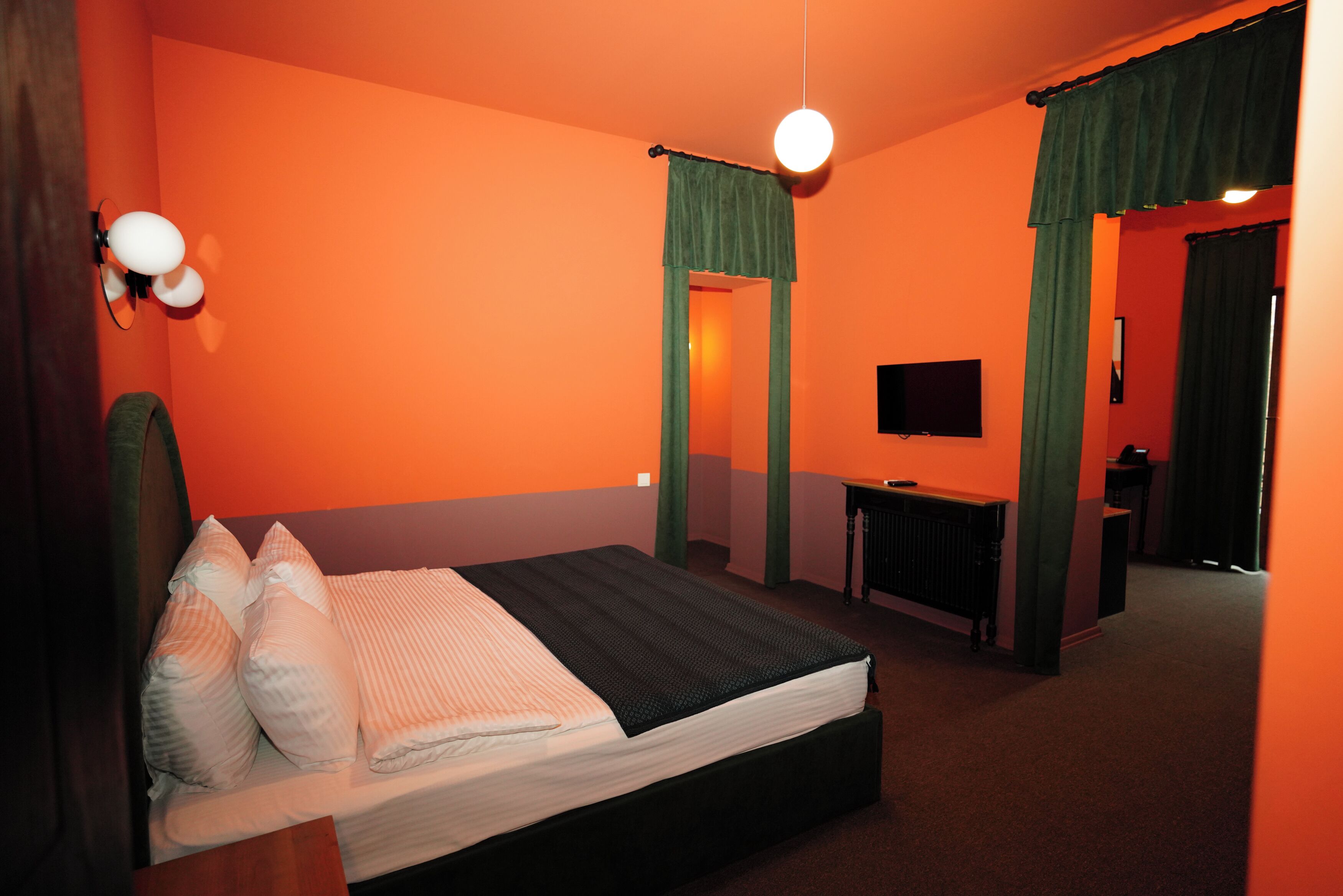 Una habitación con pared naranja, una cama con ropa de cama blanca y negra, un escritorio con una computadora y un televisor en la pared.