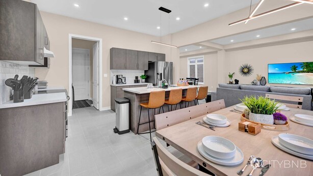 Une cuisine moderne avec des meubles gris, un coin repas avec une table en bois dressée pour quatre personnes, et un canapé gris dans le salon.