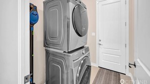 Une buanderie équipée de machines à laver et de sèche-linge superposés, d’un aspirateur et d’une porte blanche.