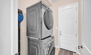 Une buanderie équipée de machines à laver et de sèche-linge superposés, d’un aspirateur et d’une porte blanche.