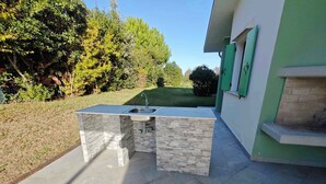 An outdoor kitchen with a sink and stone accents, located in a garden with a house in the background.