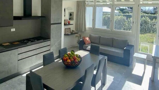 A modern kitchen with a dining area, large windows, and a view of greenery.