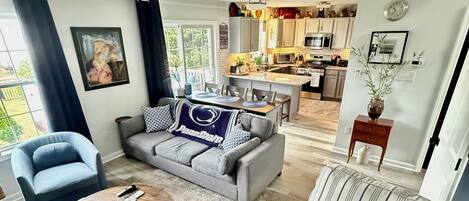 A modern living room with a grey sofa, striped armchair, and round coffee table. A kitchen is visible in the background with white cabinets and a dining area.