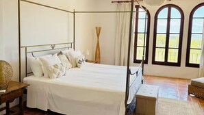 A four-poster bed with white bedding, a wooden nightstand, a woven basket, and a bench with a woven seat.