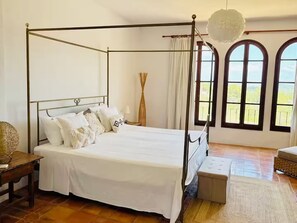 A four-poster bed with white bedding, a wooden nightstand, a woven basket, and a bench with a woven seat.