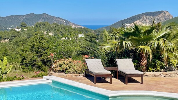 A pool area with two lounge chairs and a table, surrounded by lush greenery and mountains in the background.