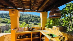 An outdoor kitchen with a view of a forested hillside, stone walls, and a tiled floor.