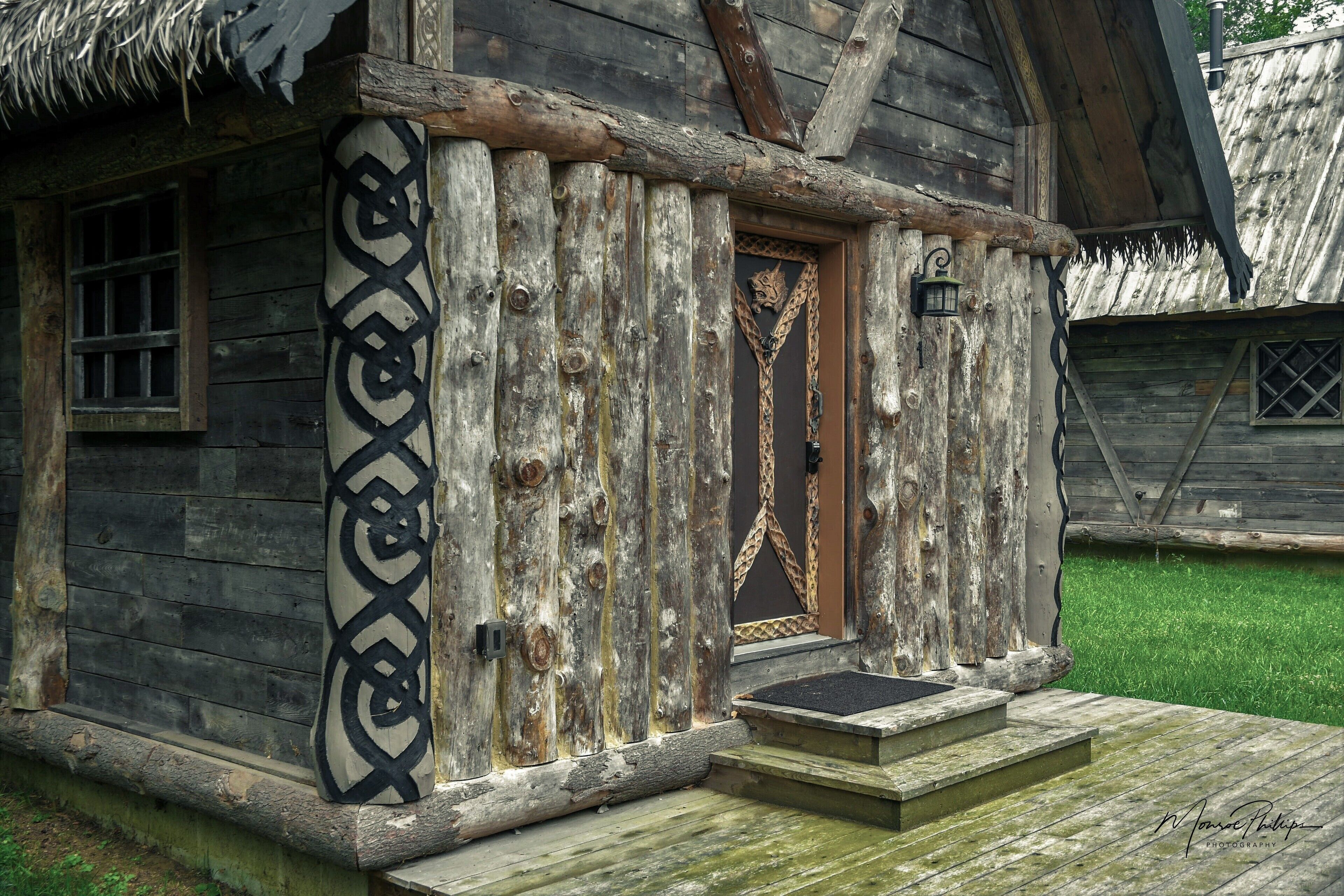 A rustic wooden cabin with a thatched roof, a wooden door with a decorative pattern, and a small porch with steps.