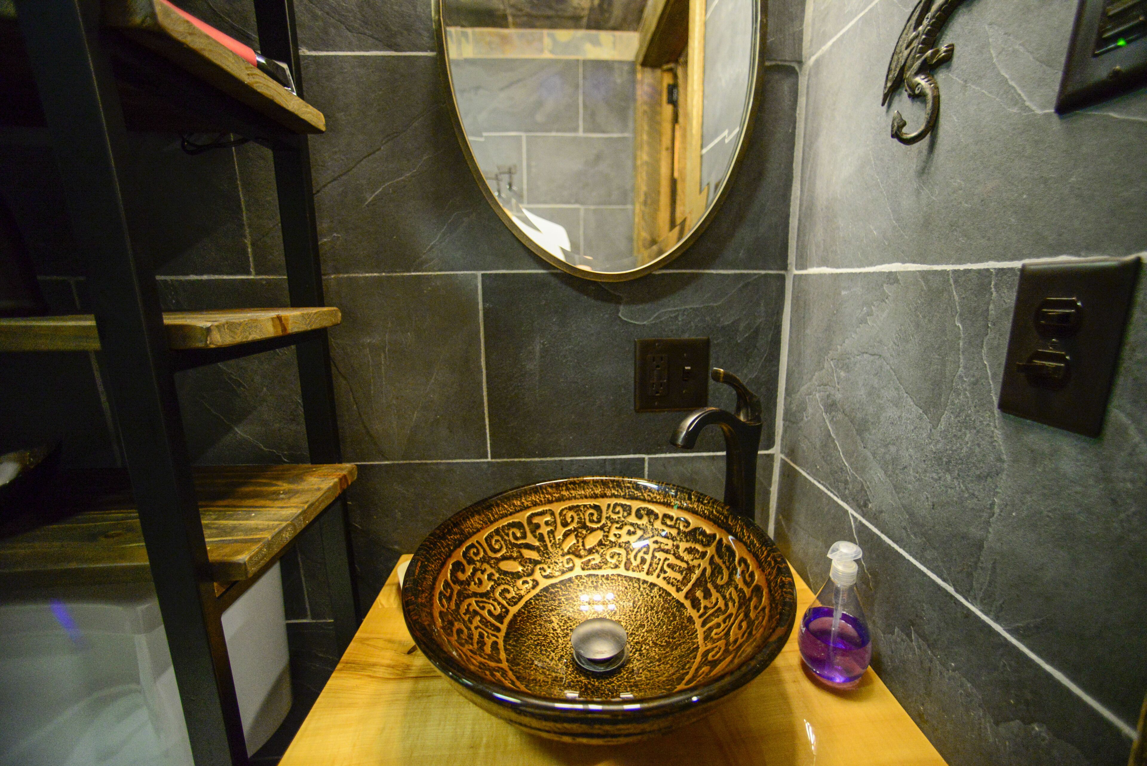 Un baño con un lavamanos grande y decorativo, un estante de madera, un espejo y una botella de gel antibacterial.