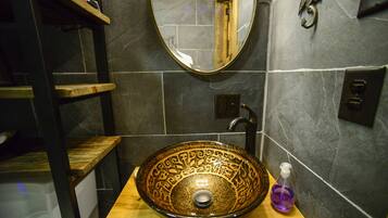Un baño con un lavamanos grande y decorativo, un estante de madera, un espejo y una botella de gel antibacterial.