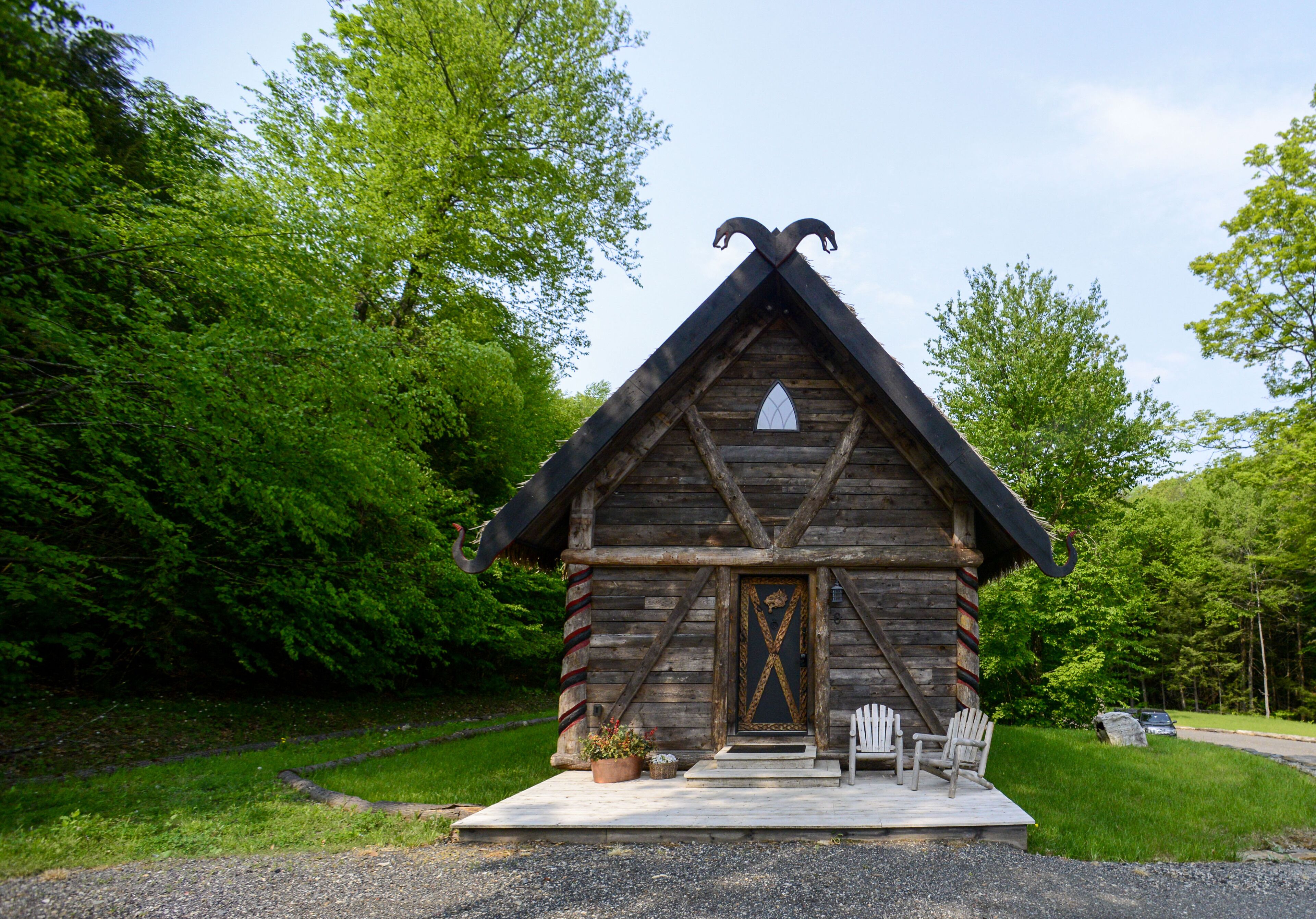 A rustic log cabin with a steeply pitched roof, a prominent door with a decorative cross, and a small porch with two Adirondack chairs.