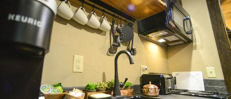 A kitchen with a black sink, a coffee maker, a toaster, and a kettle. There are also several potted plants and a wooden shelf above the sink.
