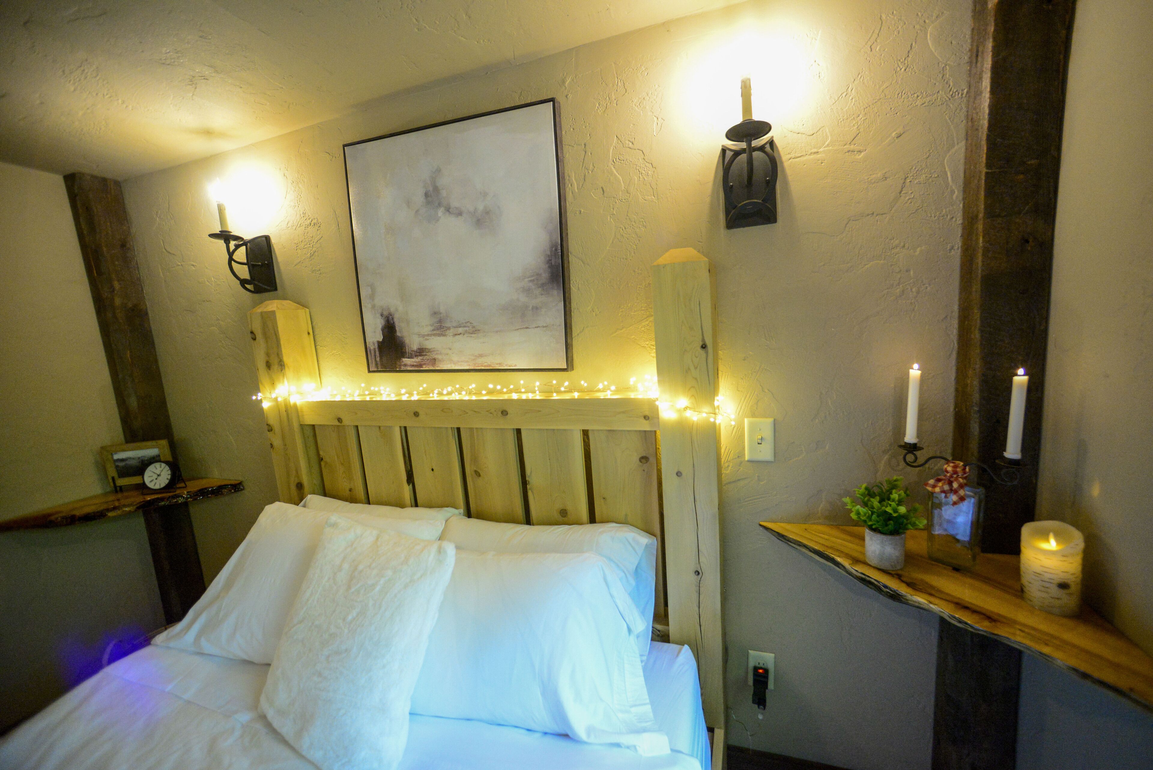 A bedroom with a wooden headboard, a bedside table with a clock, a wall-mounted painting, and string lights.