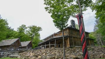 A rustic wooden building with a stone foundation, a thatched-roof structure to the left, and a prominent red and black rope sculpture.