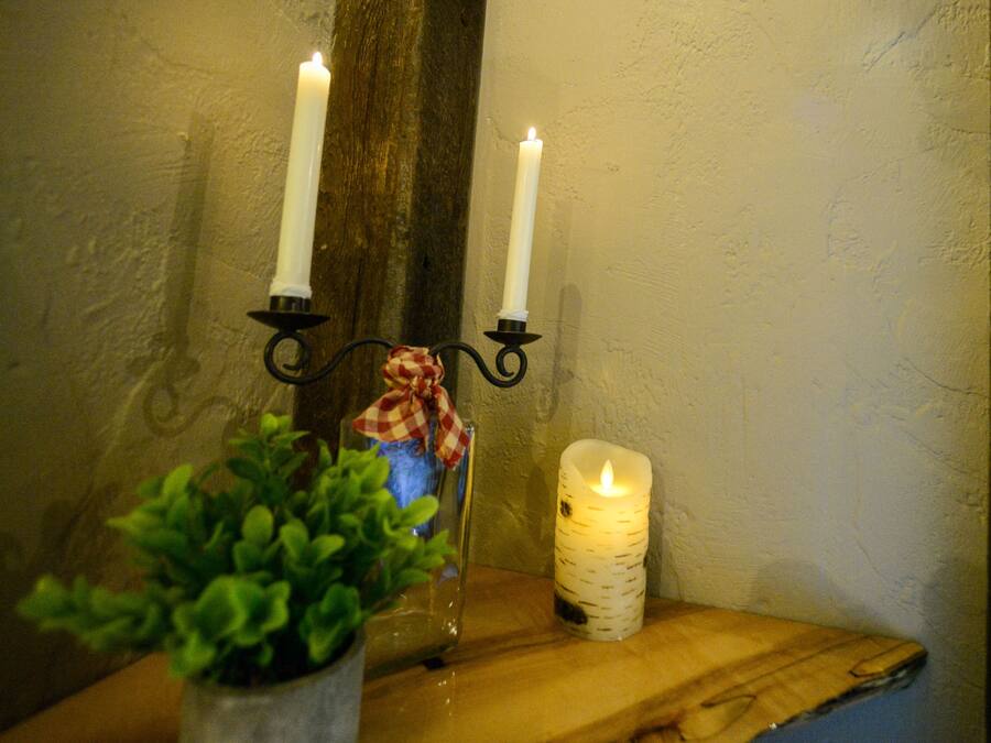 Un portavelas con dos velas encendidas y una planta pequeña sobre un estante de madera frente a una pared con textura.