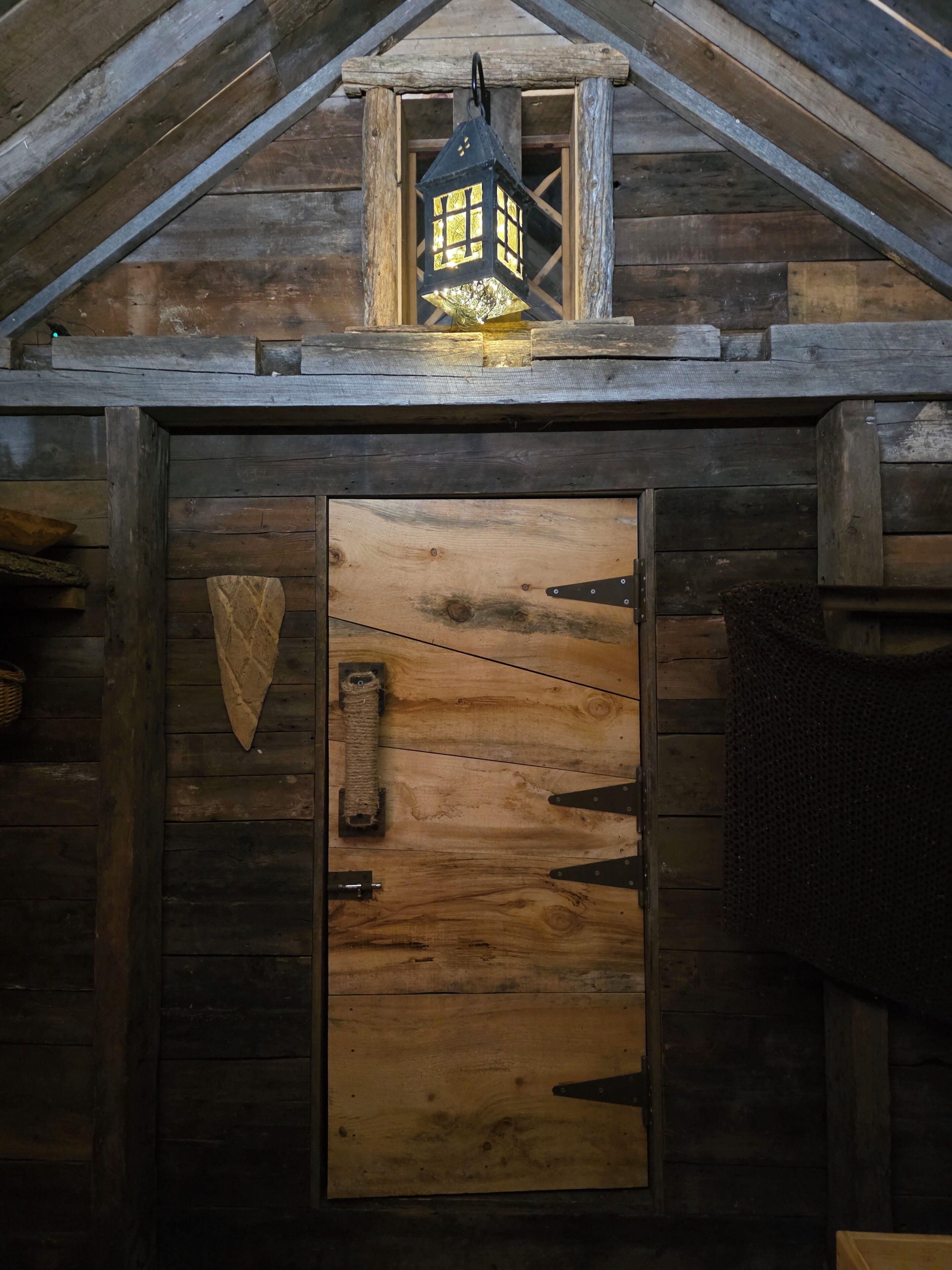 Une cabane rustique en bois, avec une porte en bois, une lanterne au-dessus de la porte et un panier tressé sur la gauche.