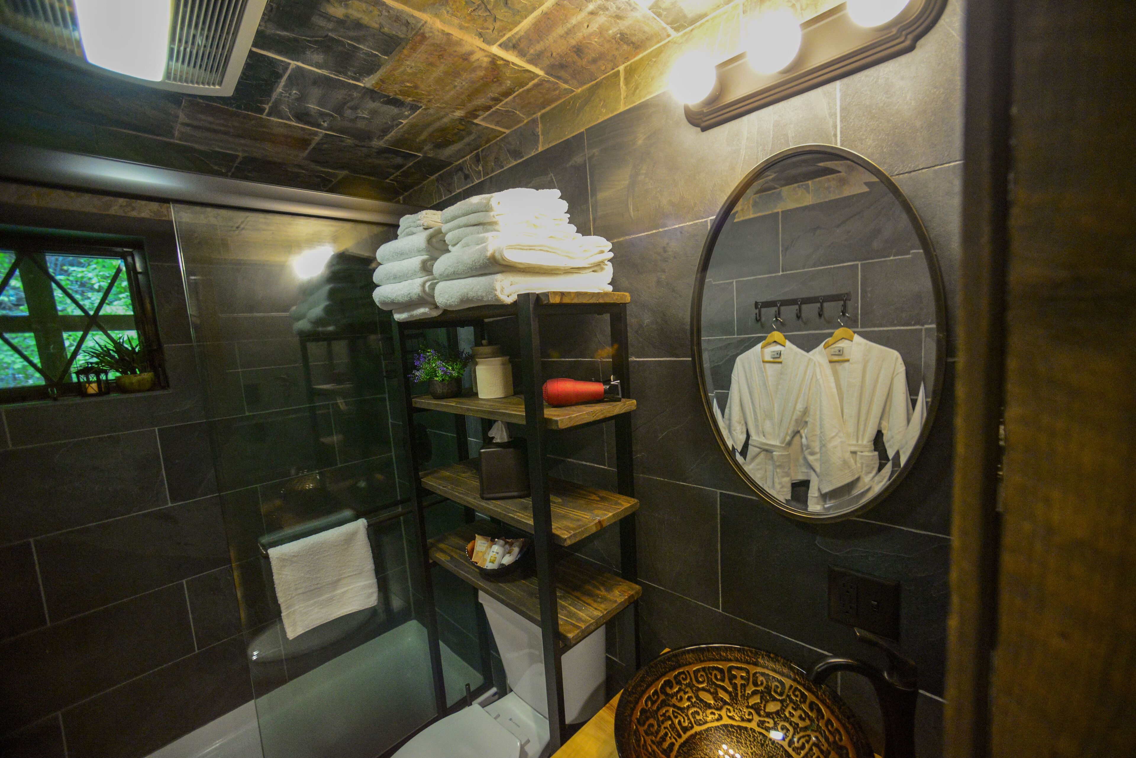 A bathroom with a glass shower enclosure, a wooden shelf with towels, a toilet, and a mirror with a towel rack.