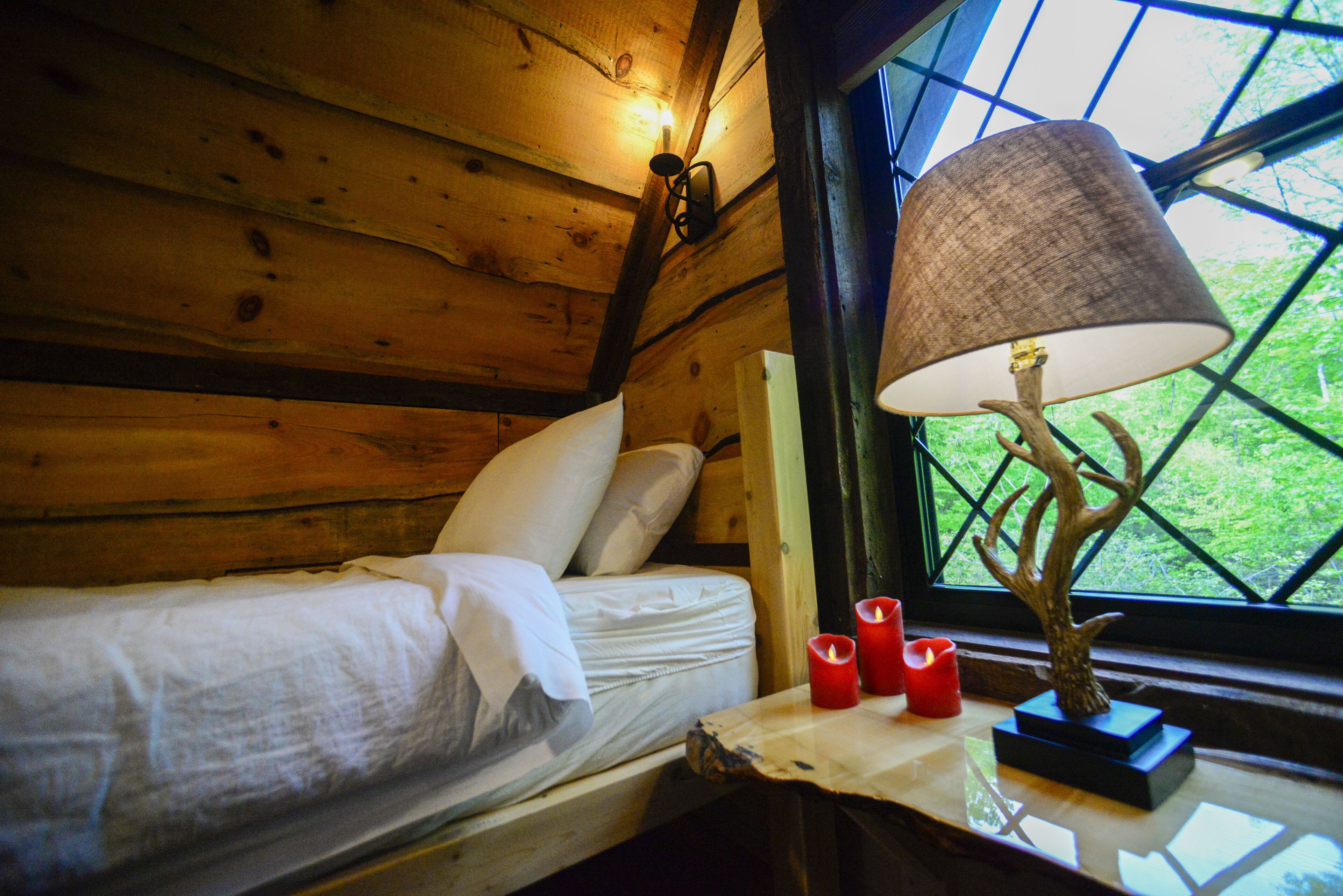 Une pièce de cabane en bois comprenant un lit, une table avec des bougies et une lampe posée sur un pied en forme d’arbre.