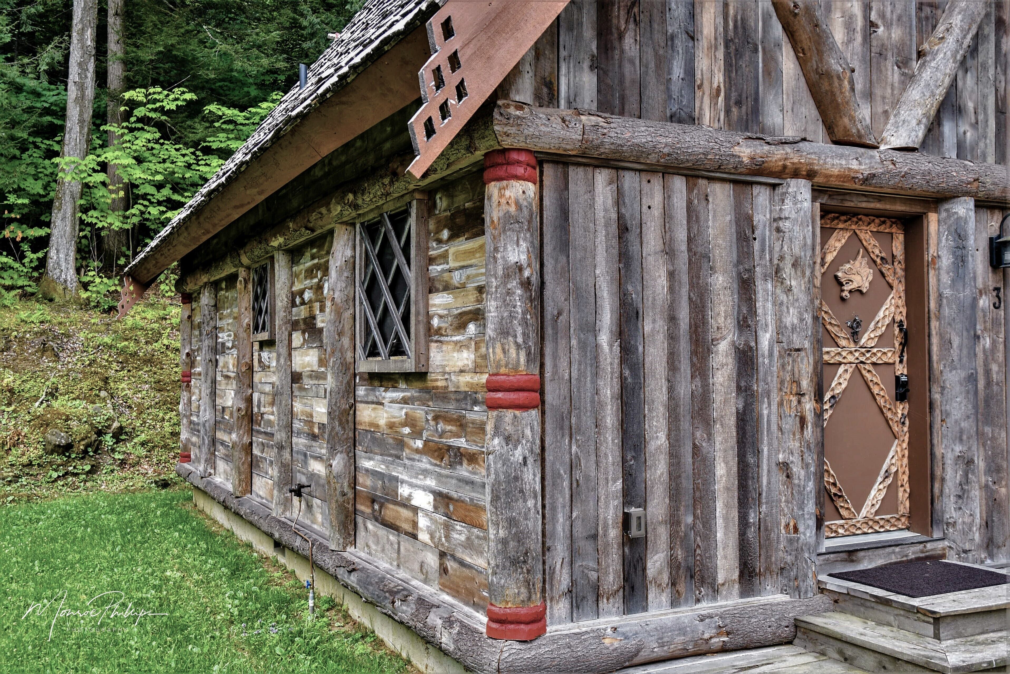 A rustic log cabin with a wooden door and windows, surrounded by greenery.