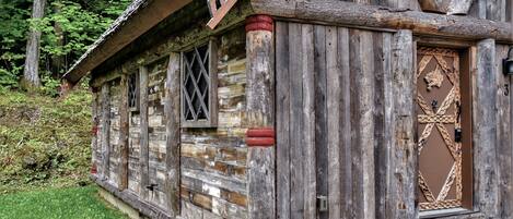 A rustic log cabin with a wooden door and windows, surrounded by greenery.