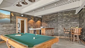 Una mesa de billar con superficie de fieltro verde, una zona de bar con mostrador de madera y un conjunto de dos sillas.
