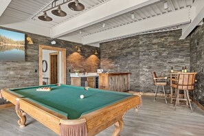 A pool table with a green felt surface, a bar area with a wooden countertop, and a seating arrangement with two chairs.