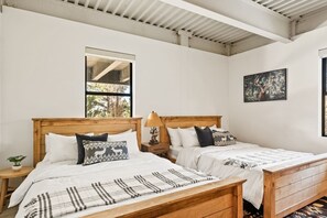 Ein Zimmer mit zwei Holzbett, einem Nachttisch, einer Lampe und einem Fenster mit Blick auf Bäume.