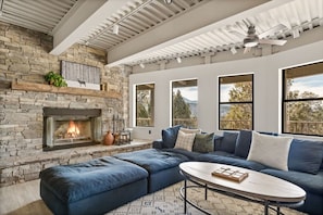 A living room with a stone fireplace, a blue sectional sofa, and large windows offering a view of trees and mountains.