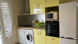 Een kleine keuken met gele kastjes, een witte koelkast, een magnetron, een wasmachine en een eethoek met een stoel en tafel.