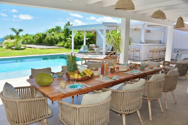 Un espace repas aménagé au bord de la piscine, avec des chaises en osier, une table en bois et une vue sur la piscine et la végétation.