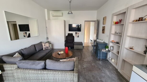Uma sala de estar moderna com um sofá, mesa de centro e televisão. Há uma estante de livros e um espaço de refeições com mesa e cadeiras.