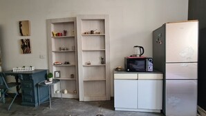 Uma cozinha compacta com micro-ondas, chaleira e frigorífico. Há uma pequena mesa de jantar com cadeiras e um armário com prateleiras.