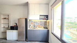 Cozinha compacta com geladeira, micro-ondas e pia. Uma janela oferece vista para o horizonte urbano.