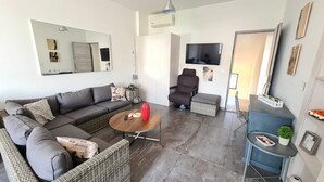 Uma sala de estar moderna com sofá, poltrona e mesa de centro. Há uma televisão na parede, uma mesinha lateral e um espelho.