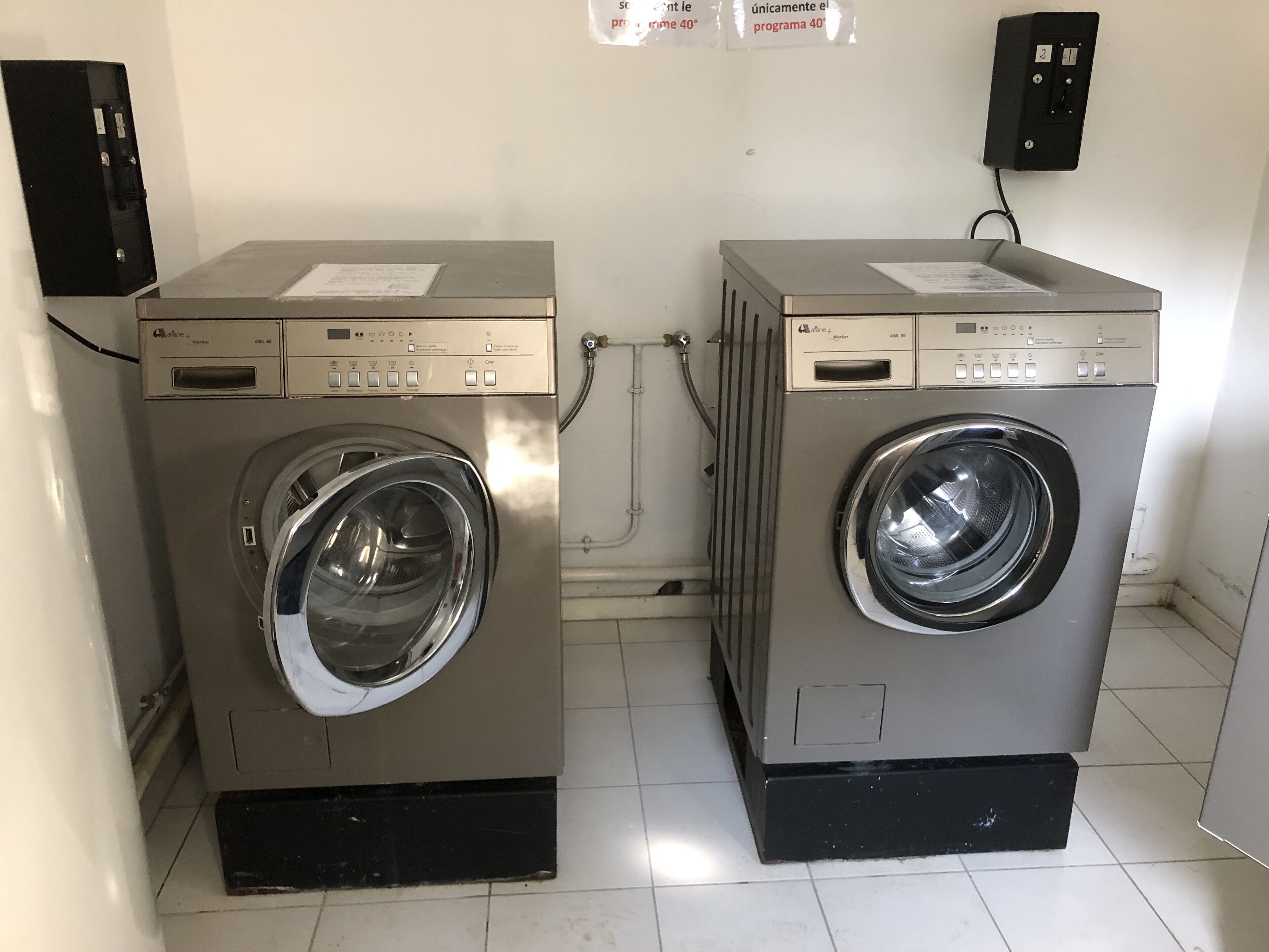 Deux machines à laver à chargement frontal se trouvent dans une pièce dont le sol est recouvert de carreaux blancs et dont un des murs est uni.