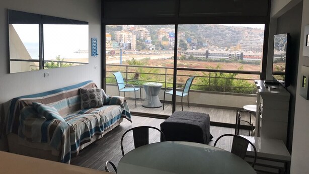 A modern living room with a sofa, chairs, a dining table, and a balcony with a view of a cityscape.