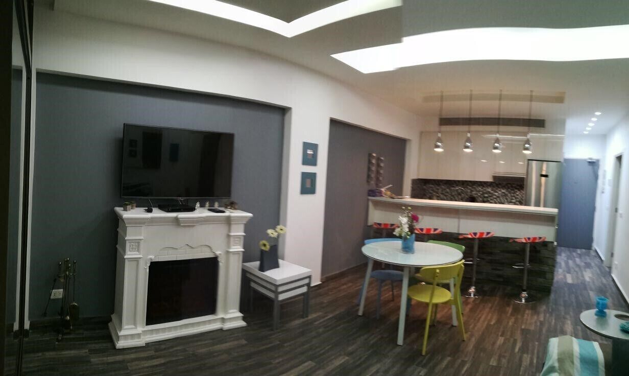 A modern living room with a fireplace, a television, a dining area with a round table and chairs, and a kitchen area with a countertop and hanging lights.