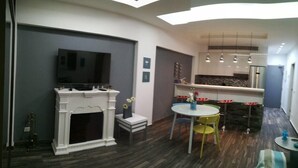 A modern living room with a fireplace, a television, a dining area with a round table and chairs, and a kitchen area with a countertop and hanging lights.