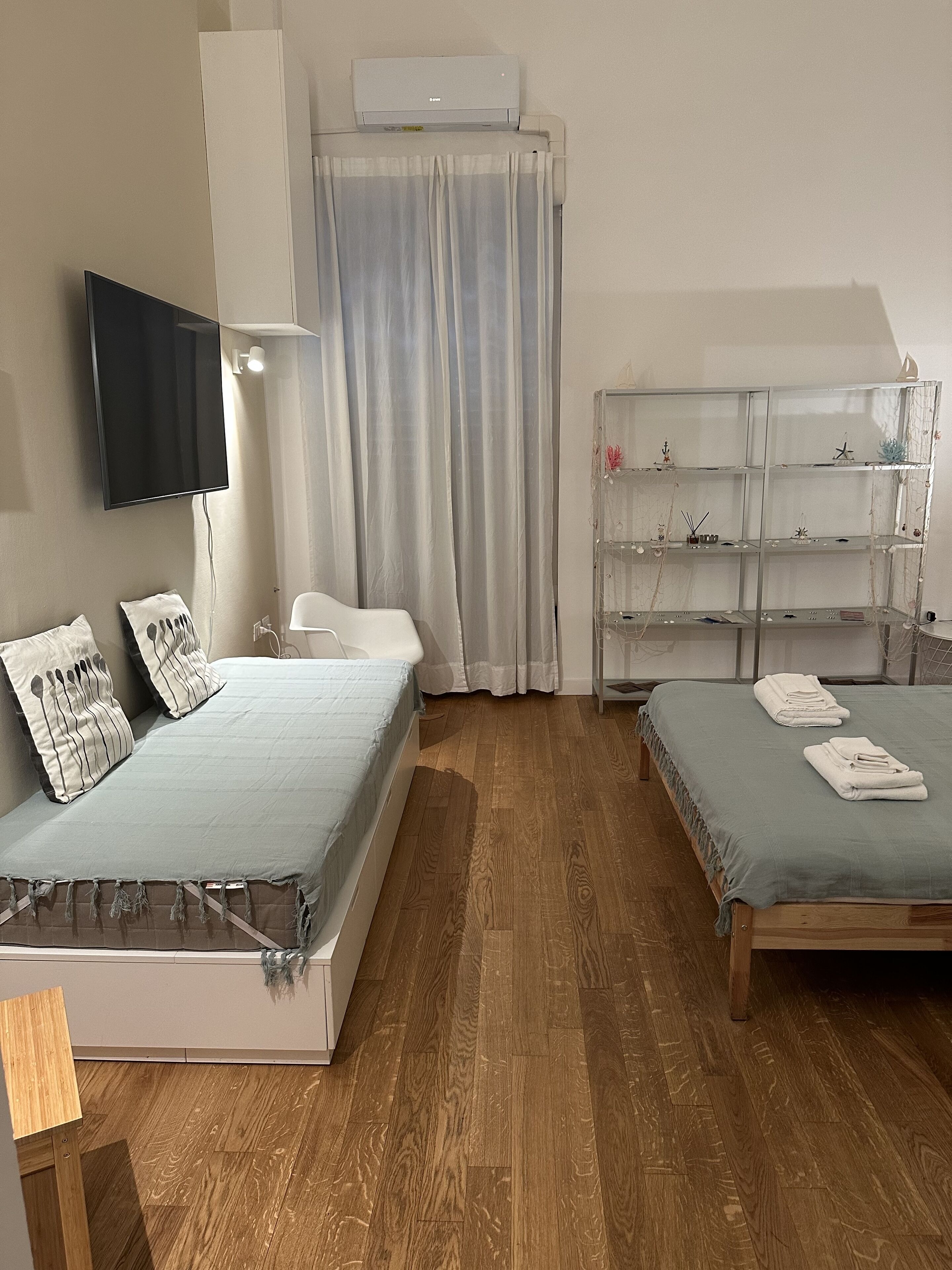 A bedroom with two beds, a TV, a shelf with decorative items, and a window with curtains.
