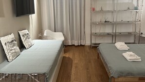 Et soveværelse med to senge, et fjernsyn, en reol med pynteting og et vindue med gardiner.