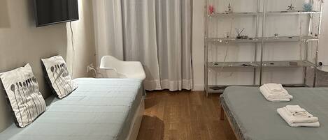 A bedroom with two beds, a TV, a shelf with decorative items, and a window with curtains.