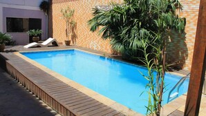 Ein rechteckiger Swimmingpool mit Holzdeckrand, umgeben von einer Backsteinmauer mit dekorativem Muster. Liegestühle und Topfpflanzen befinden sich rund um den Pool.