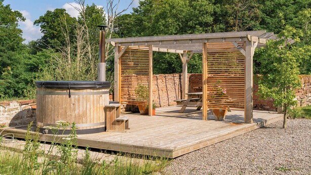 Een houten terras buiten met een jacuzzi, een pergola en een stenen muur.