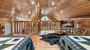 Eine Holzhütte mit Holzwänden und -decke, zwei Betten mit gemusterten Quilts, einem großen schwarzen Lederteppich, einer Holzbank und Blick auf einen Wald durch ein Fenster.