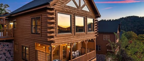 A log cabin with a balcony, stone foundation, and a view of the surrounding landscape.
