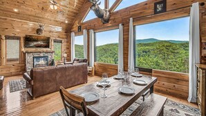 Interior de una cabaña de madera con una mesa de comedor, una chimenea, un sofá de cuero y un ventanal con vistas a un bosque.