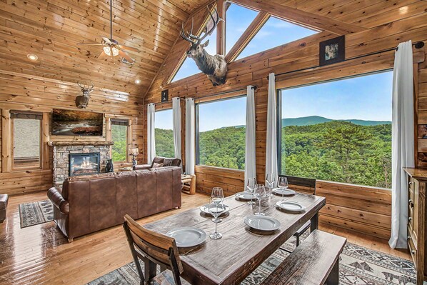 A wooden cabin interior with a dining table set, a fireplace, a leather sofa, and a large window overlooking a forest. - Epic Mountain Views, Hot Tub, Sleeps 10, Pool Table, Arcade, Foosball, Fast WiFi (Sevierville)