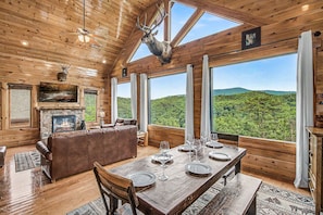 A wooden cabin interior with a dining table set, a fireplace, a leather sofa, and a large window overlooking a forest.